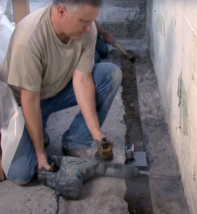 Worker using a power jackhammer to break concrete in a narrow trench beside a wall